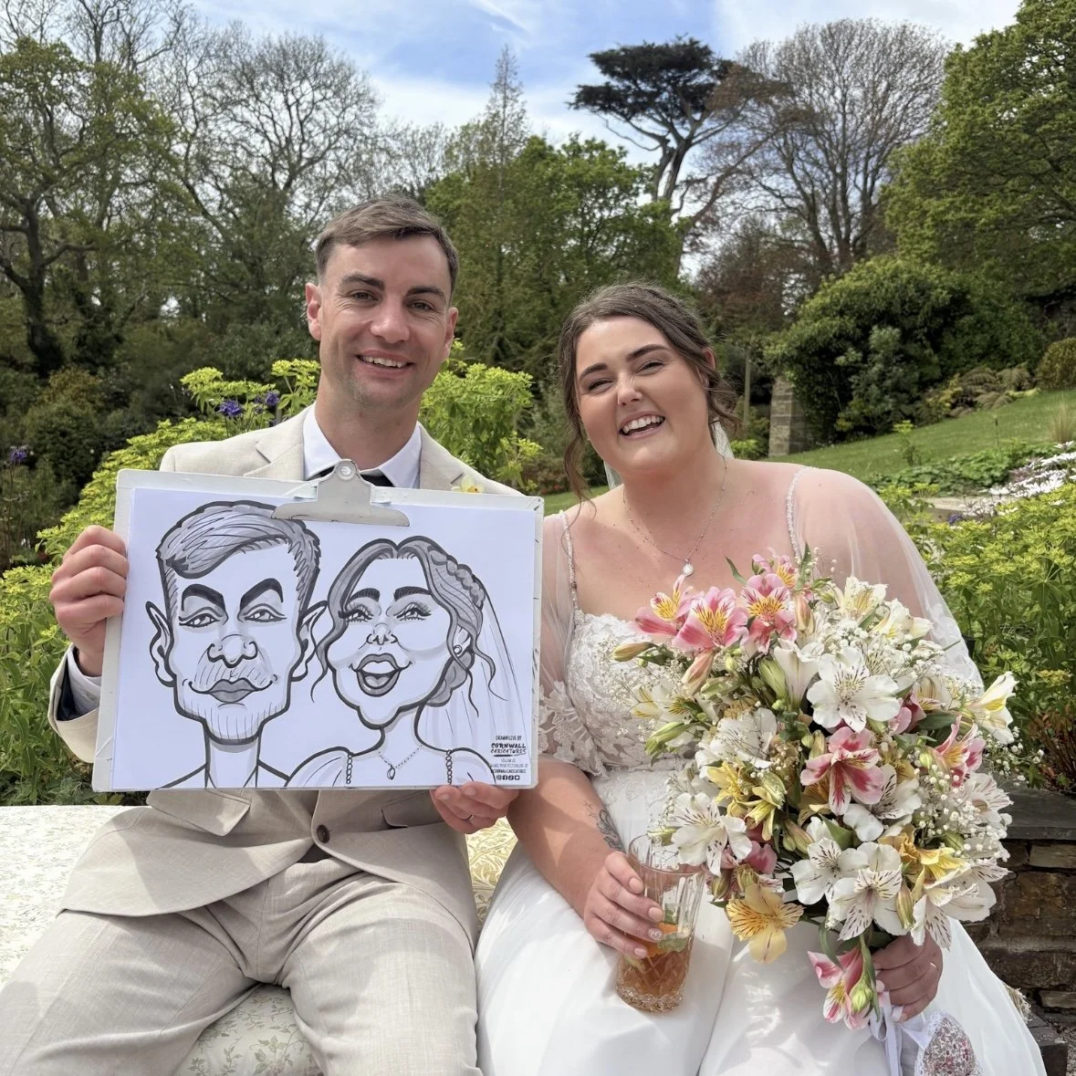 A smiling bride and groom sit outdoors in a garden, the bride holding a large bouquet of white and pink flowers, the groom holding a caricature drawing of them in a frame.