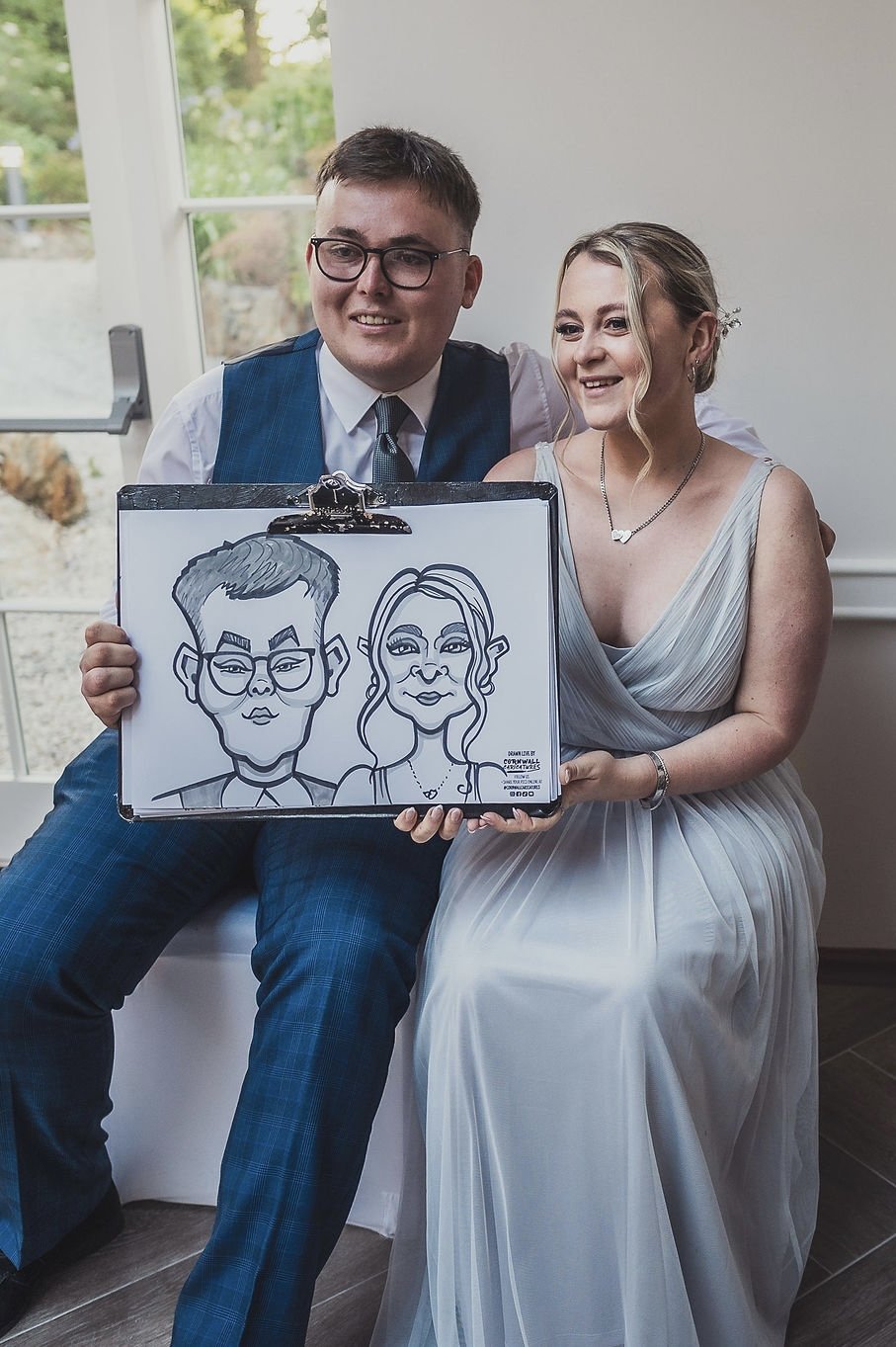 A couple dressed in wedding attire sitting indoors near a window, holding a caricature drawing of themselves on a clipboard.