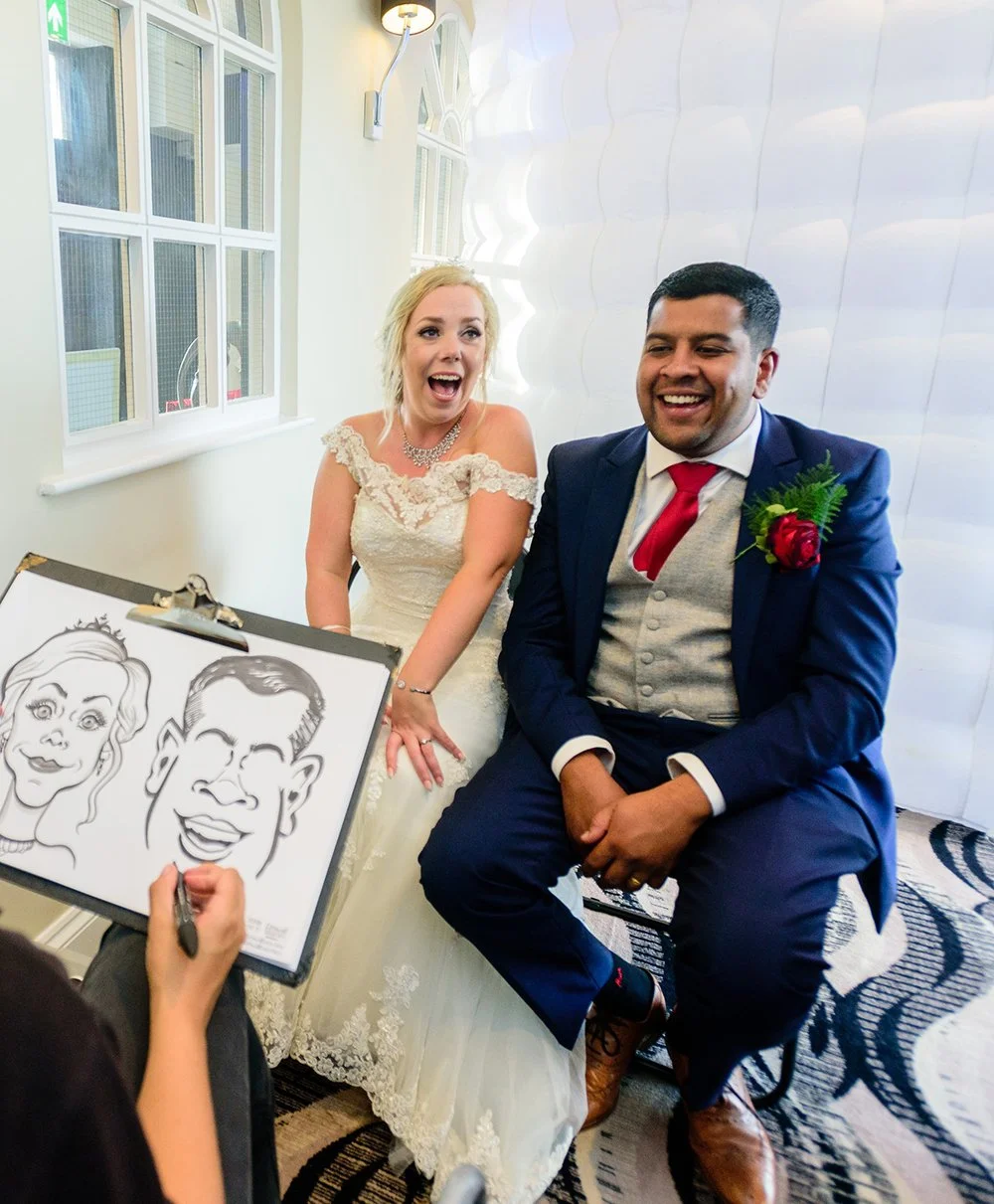 A bride and groom sitting together, laughing during their wedding, with caricature artist drawing their portraits.