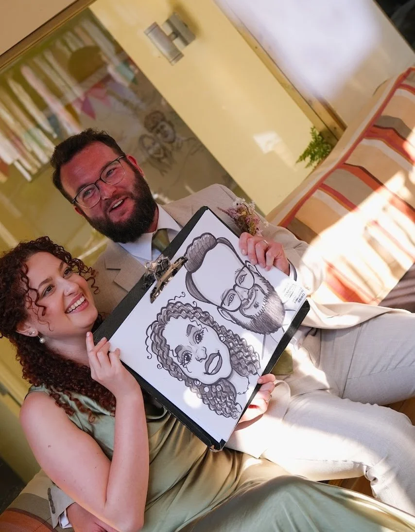 A smiling woman with curly hair and pearl earrings holding a caricature sketch of herself and a man with a beard and glasses, in a cozy, warmly lit restaurant or cafe.