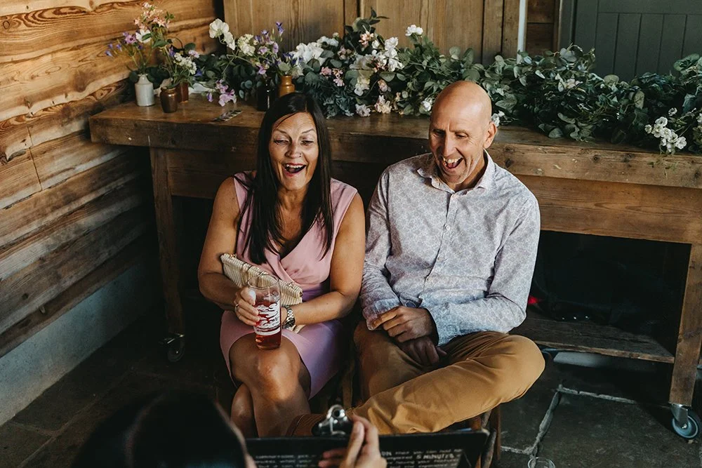 A woman and man sitting on a bench, smiling and laughing, with the woman holding a drink, in a rustic setting with wooden paneling and floral decorations in the background.