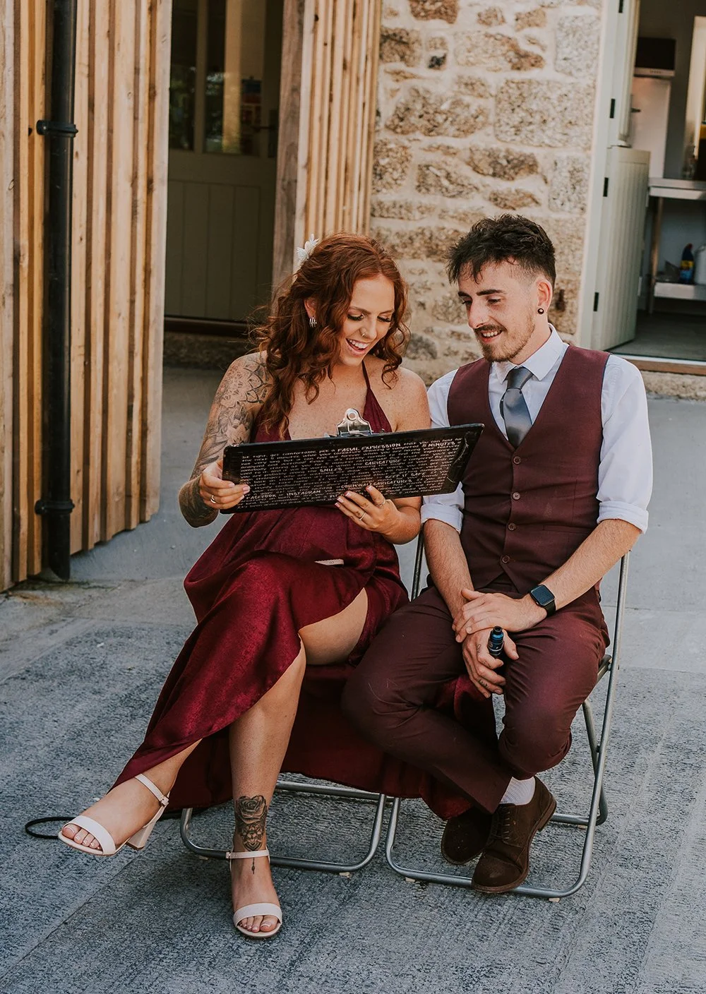 A woman and a man sitting together outdoors, looking at a black clipboard holding a caricature, smiling and sharing a happy moment.