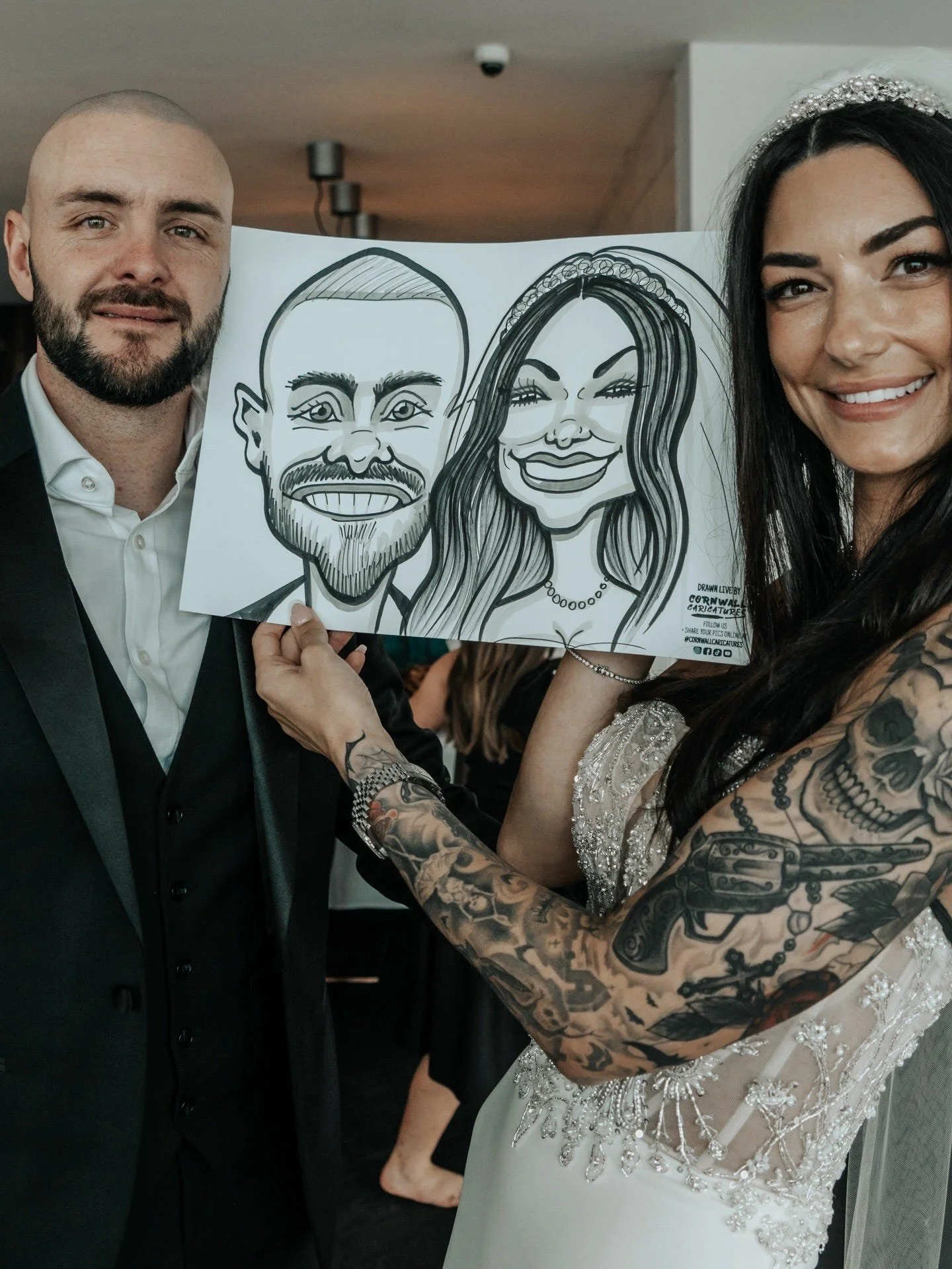 A bride with dark hair and tattoos on her arms is showing a caricature drawing of herself and a groom in a tuxedo. The groom has a beard and short hair. They are smiling at a celebration or wedding.