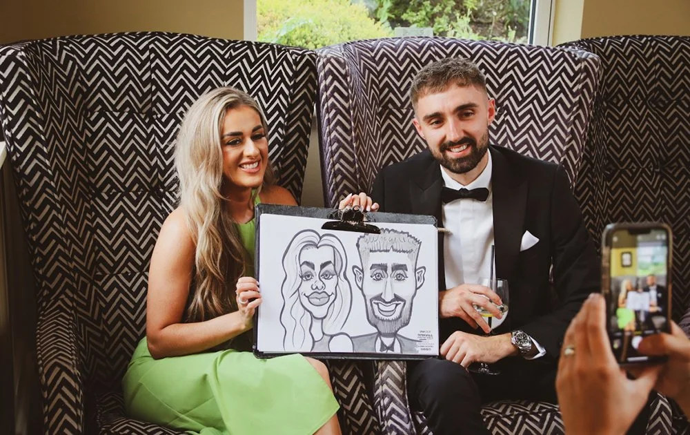 A smiling woman in a green dress and a man in a tuxedo sitting on a patterned booth, holding a caricature of themselves, while someone takes a photo.