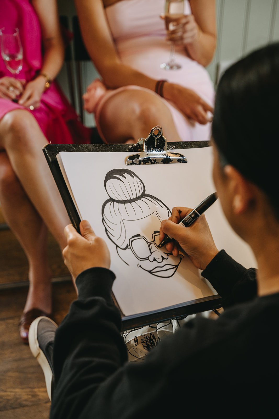 Artist drawing a black and white cartoon of a woman with sunglasses and hair in a bun on a clipboard in a social setting with 2 women in pink and peach dresses sitting in the background.