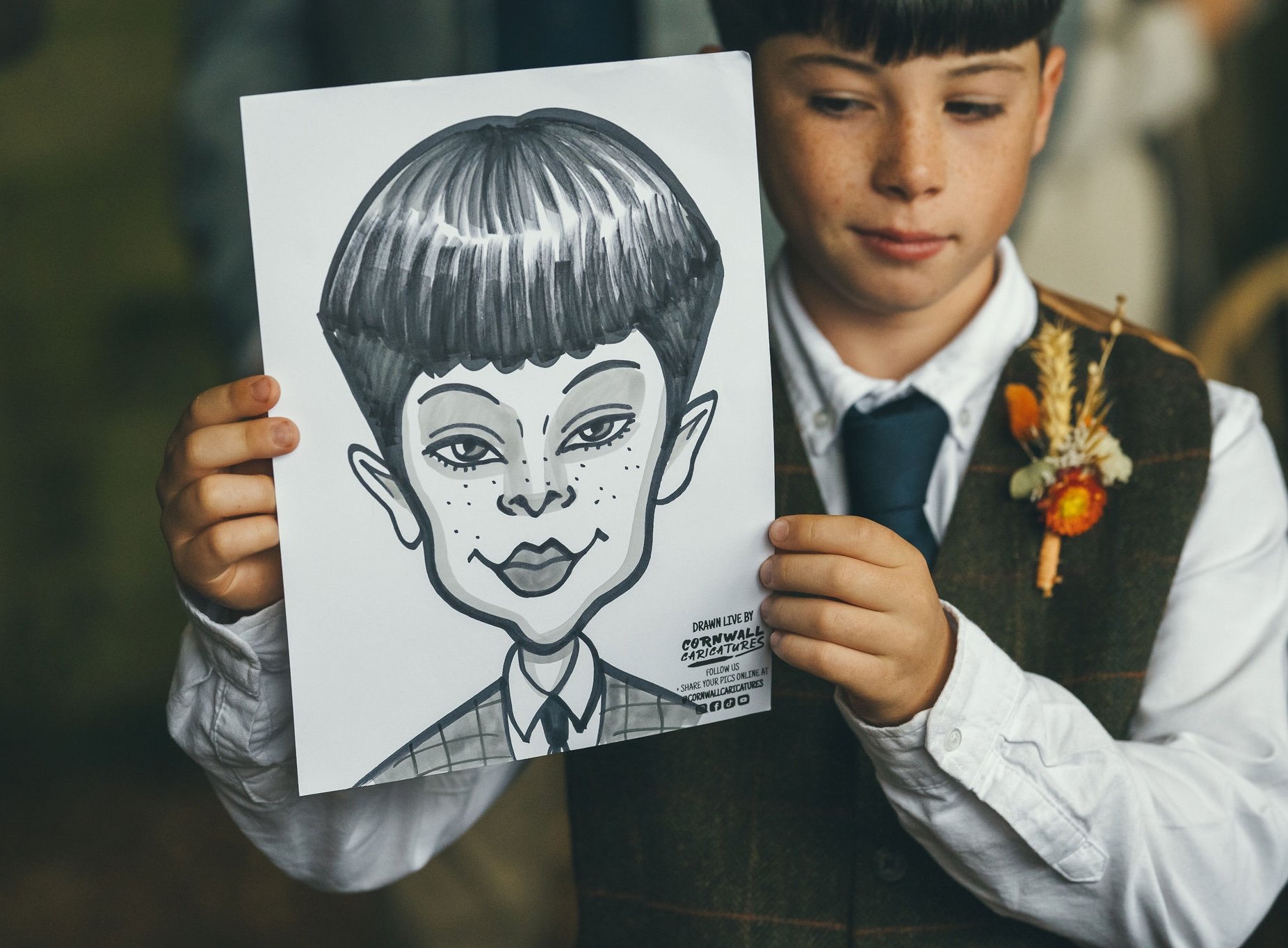 A young boy in a waistcoat holding a black and white caricature drawing of himself on a piece of paper.