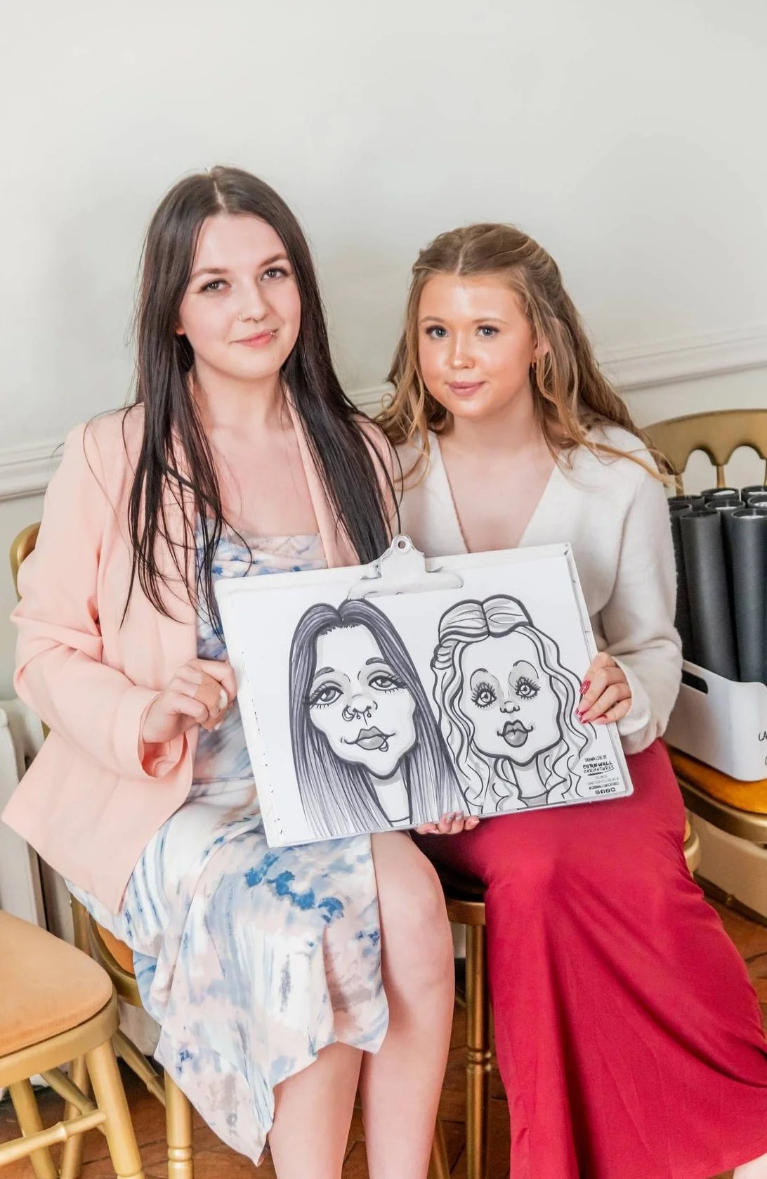 Two women sitting on chairs, holding a board with caricature drawings of their faces, in an indoor setting.