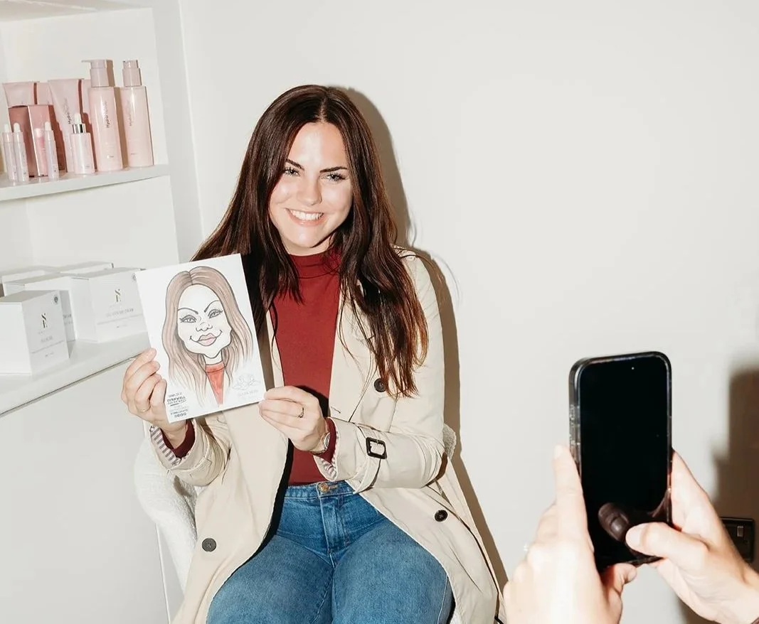 A woman with long brown hair smiling and holding a caricature portrait of herself. She is sitting in a well-lit room with a white wall and shelves of pink skincare products in the background. Someone is taking her photo with a smartphone.