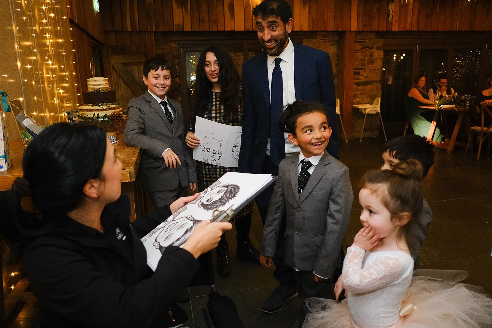 Children and adults gathered indoors at a celebration, with one woman drawing caricatures of the children, who are dressed in formal attire, in a warmly decorated room with fairy lights.