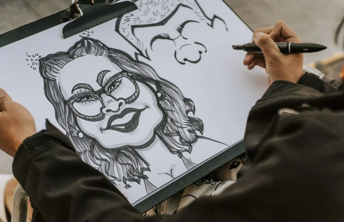 A person drawing two caricature portraits on a clipboard, one of a woman with glasses and curly hair, and another of a person with pointy ears and nose.