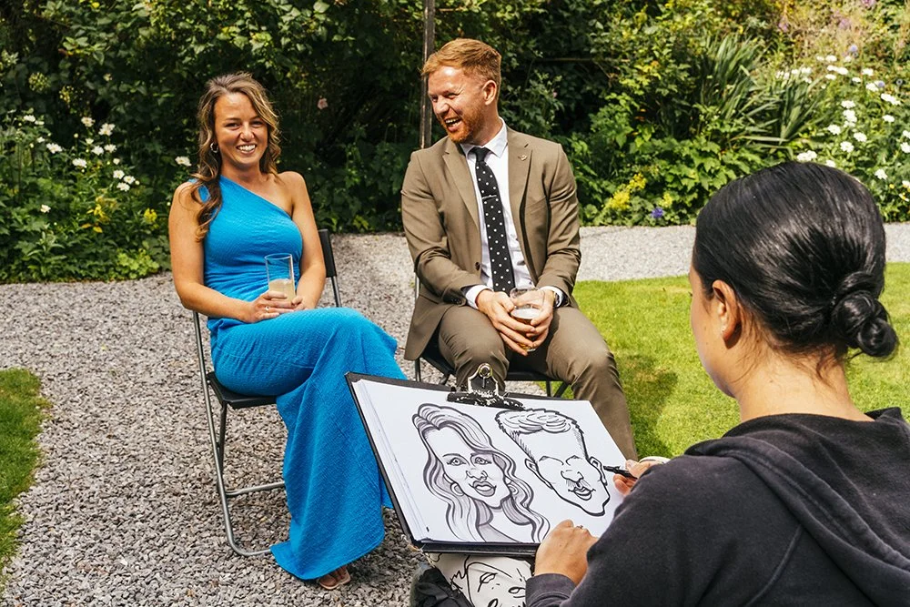 A woman and a man sitting outdoors, smiling and holding drinks, while an artist draws caricatures of them, with greenery in the background.