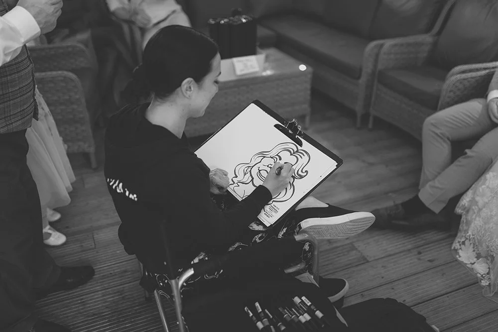 A woman sitting in a chair drawing a portrait of a woman with long hair on a clipboard in a cozy lounge.