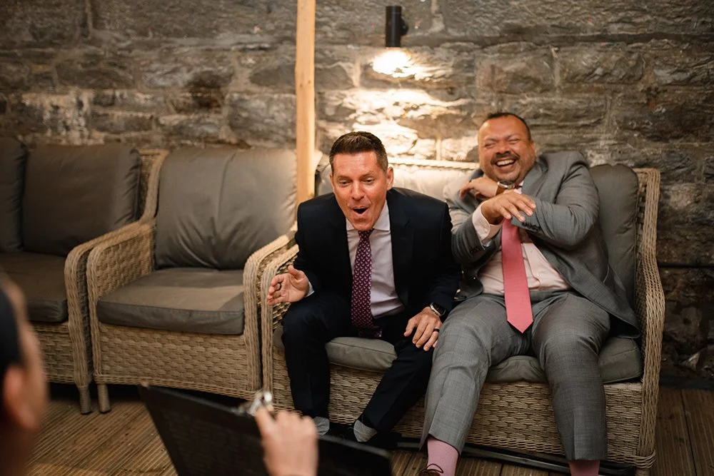 Two men in suits laughing and reacting in a cozy, rustic lounge with stone walls and wicker furniture.