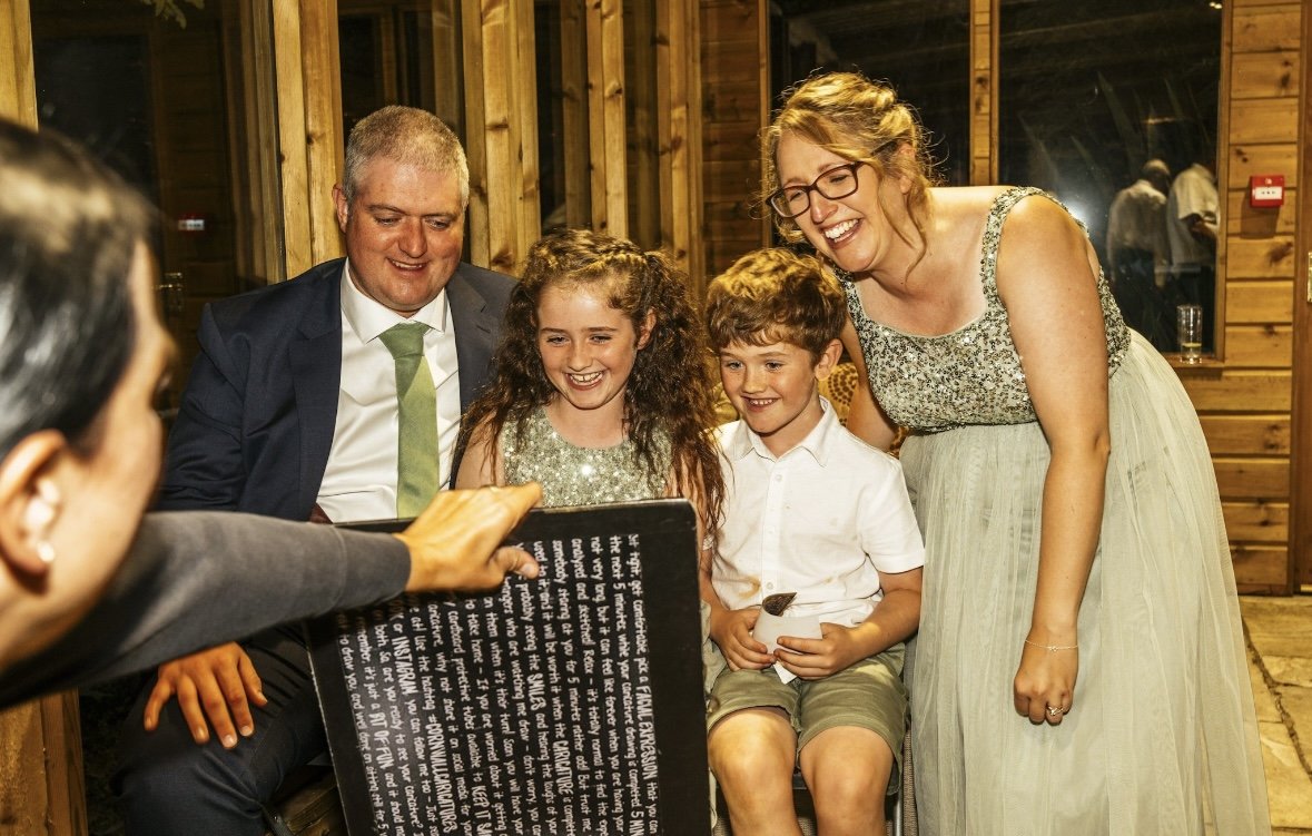 A group of five people, including two adults and three children, gathered together indoors in a wooden room, looking at a caricature and laughing.