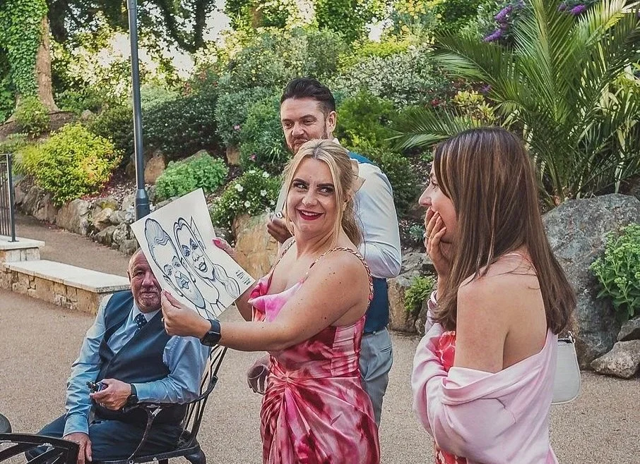 A group of four people outdoors, with a woman holding a caricature drawing of two faces. The group appears happy and engaged, surrounded by greenery and rocks.