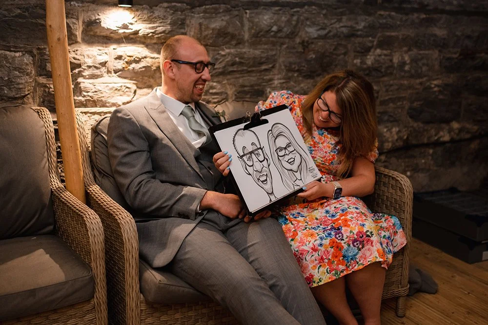 A man and a woman sitting on a wicker sofa, sharing a humorous caricature drawing of themselves on a clipboard, with both smiling and laughing. The background features a stone wall.
