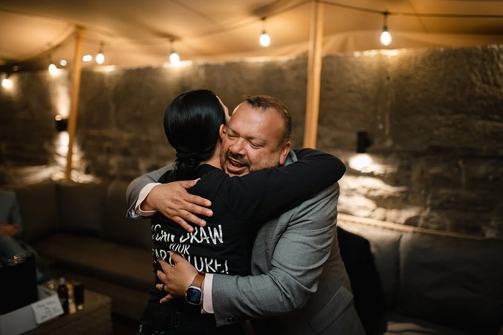 Two people hugging warmly in a cozy indoor setting with warm lighting and stone walls.