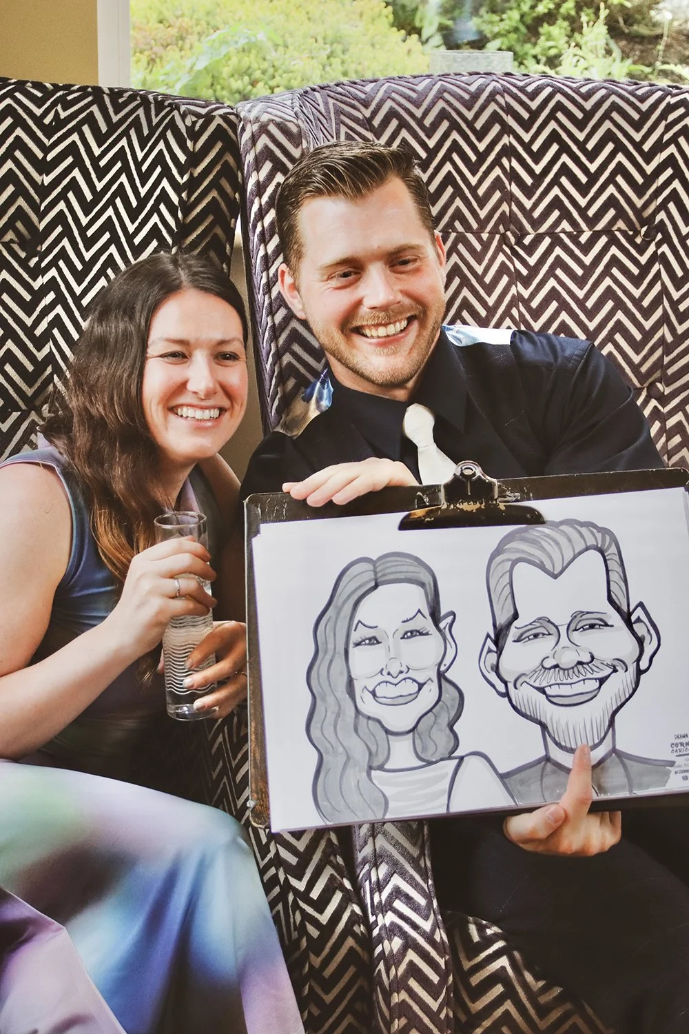 A woman and a man smiling, holding a glass of champagne and a caricature drawing of themselves, sitting in chairs with patterned backdrops and a window showing greenery outside.