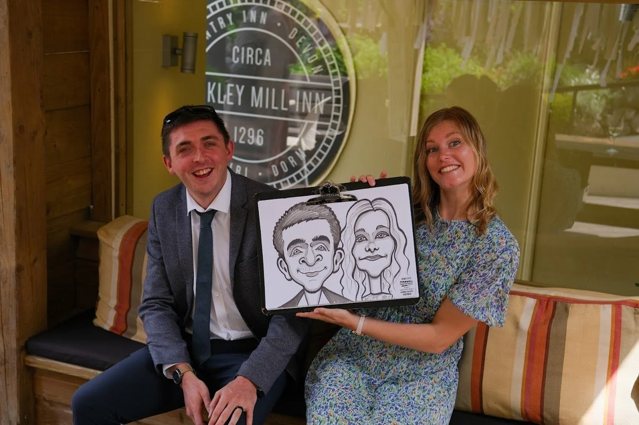 A man in a suit and a woman in a floral dress sitting on a bench, smiling, with the woman holding a caricature drawing of a man and a woman. They are indoors with a sign for Bickley Mill Inn visible in the background.
