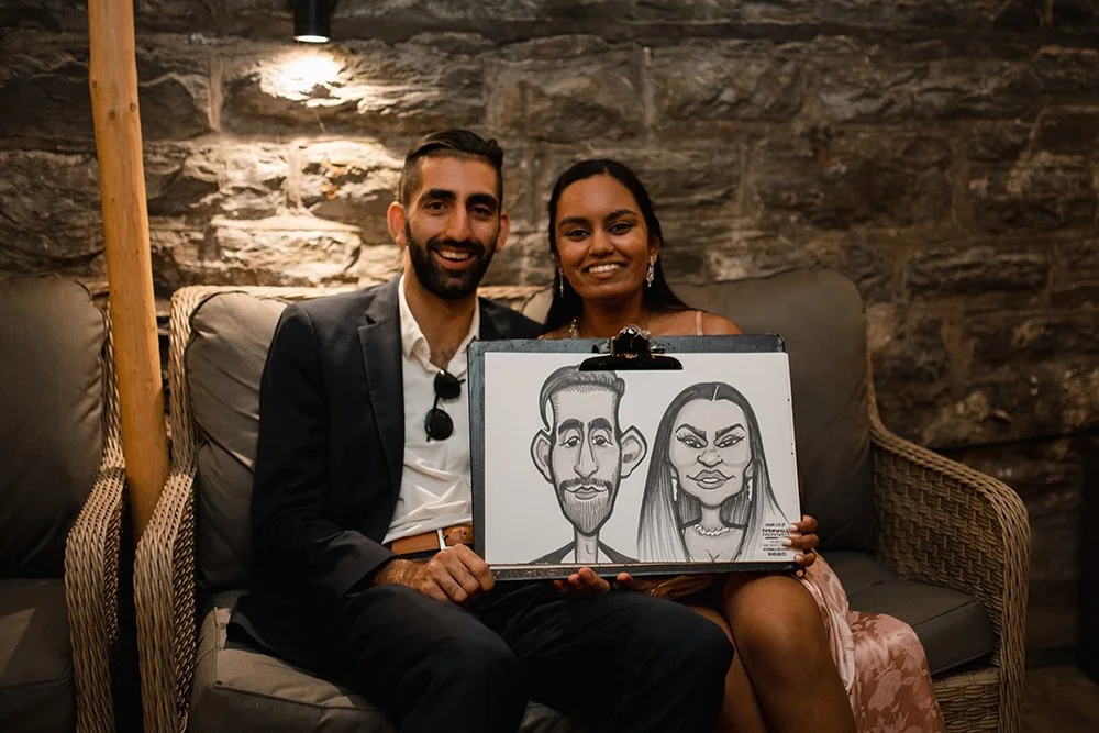 A smiling man in a dark suit and woman in a pink dress sitting on a wicker sofa in a rustic setting, holding a caricature drawing of themselves.