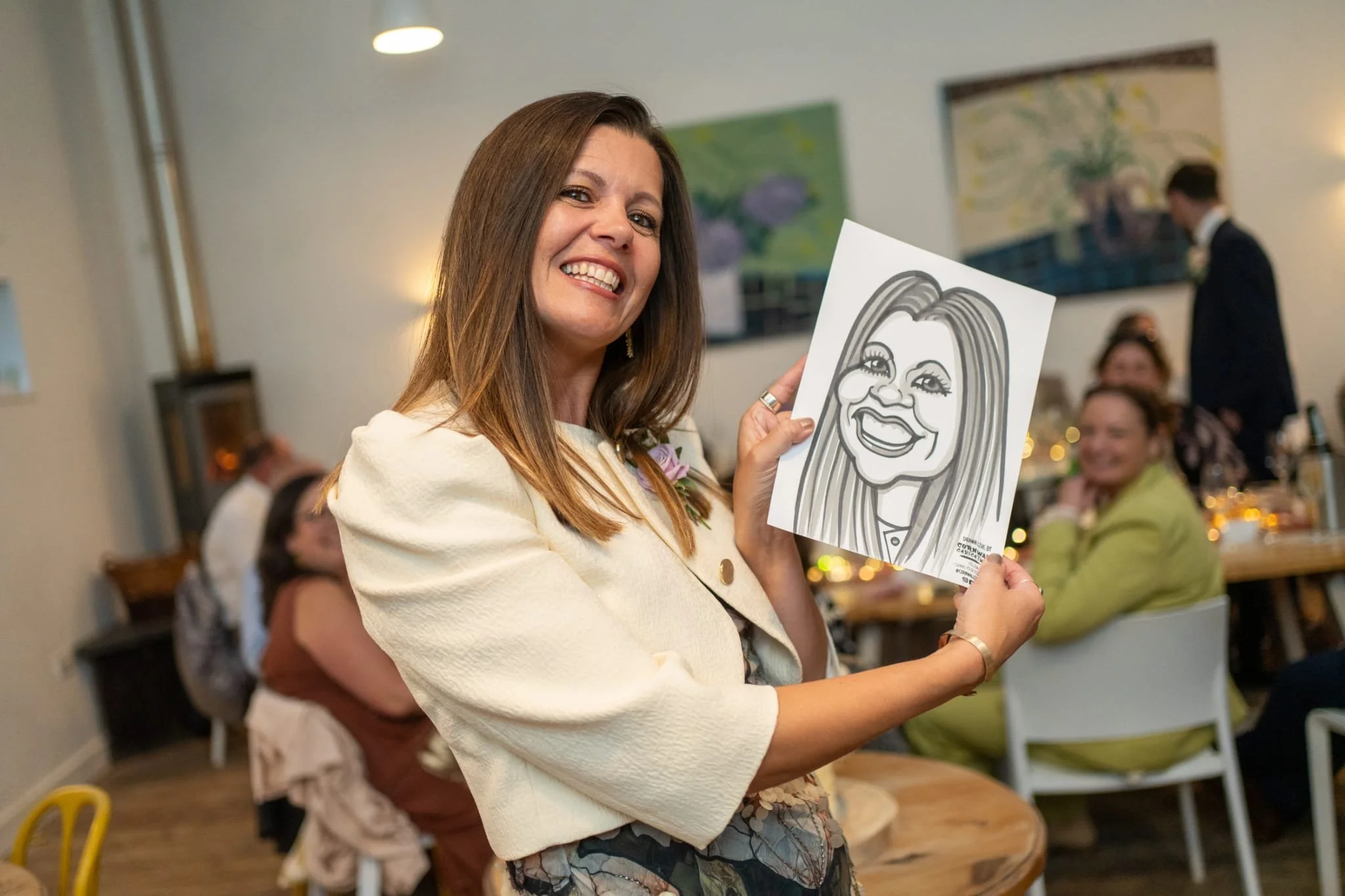 A woman in a white jacket smiling and holding a caricature drawing of herself at a gathering or celebration with people in the background.