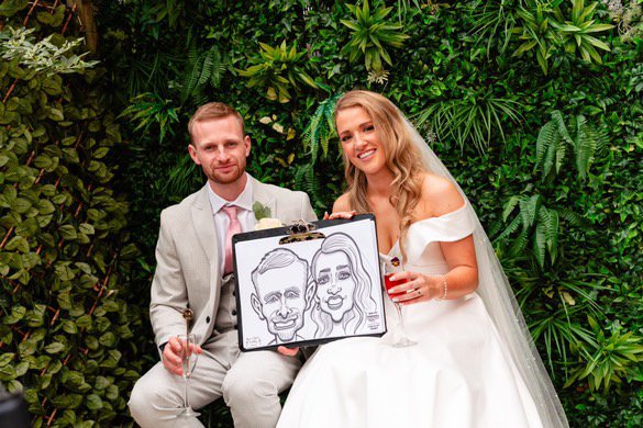 Bride and groom sitting in front of a green leafy wall, holding a drawing of themselves.