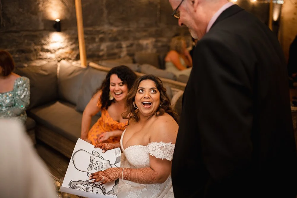 A bride with henna tattoos on her hands and wearing a white off-the-shoulder wedding dress, looking surprised and happy while holding a caricature portrait at a wedding reception. She is surrounded by friends, including one in an orange dress, in a w