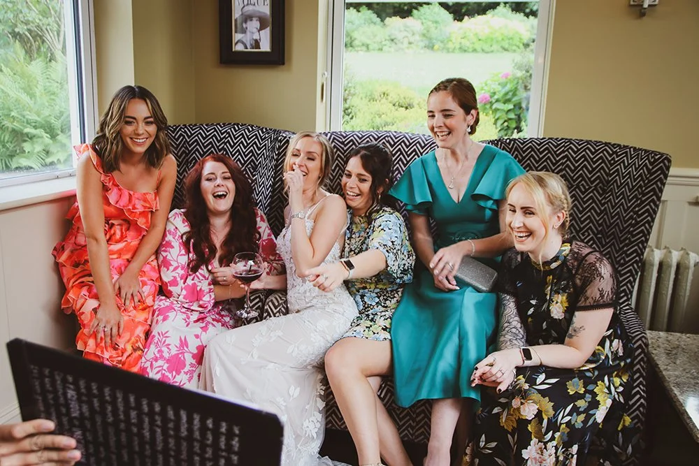 A group of six women sitting on a patterned couch, laughing and enjoying each other's company, dressed in colorful, elegant dresses, with a window showing greenery in the background.