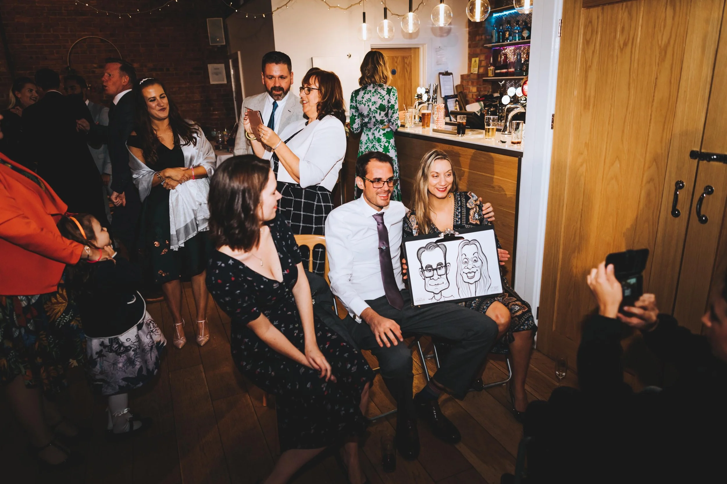 A group of people at a social gathering, some sitting and some standing, in a warmly lit bar or restaurant with wood and brick decor. One person in the center holds caricature drawings of a man and woman.
