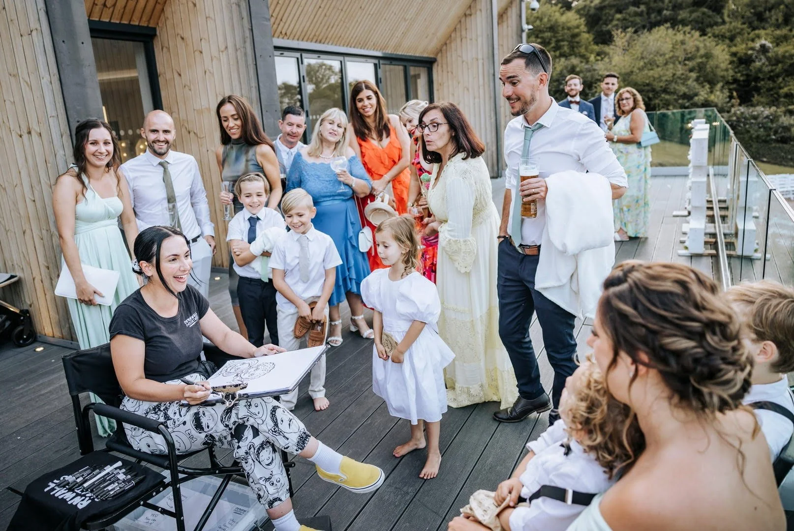 Group of people gathered on a rooftop deck, watching an artist draw caricatures of children, with friends and family smiling and socializing.