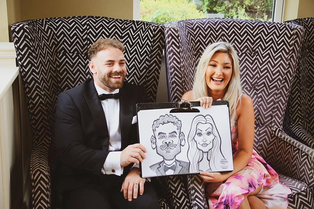 A man in a tuxedo and a woman in a colorful dress sit inside a booth, holding a caricature drawing of themselves. They are smiling and appear to be at a festive event.