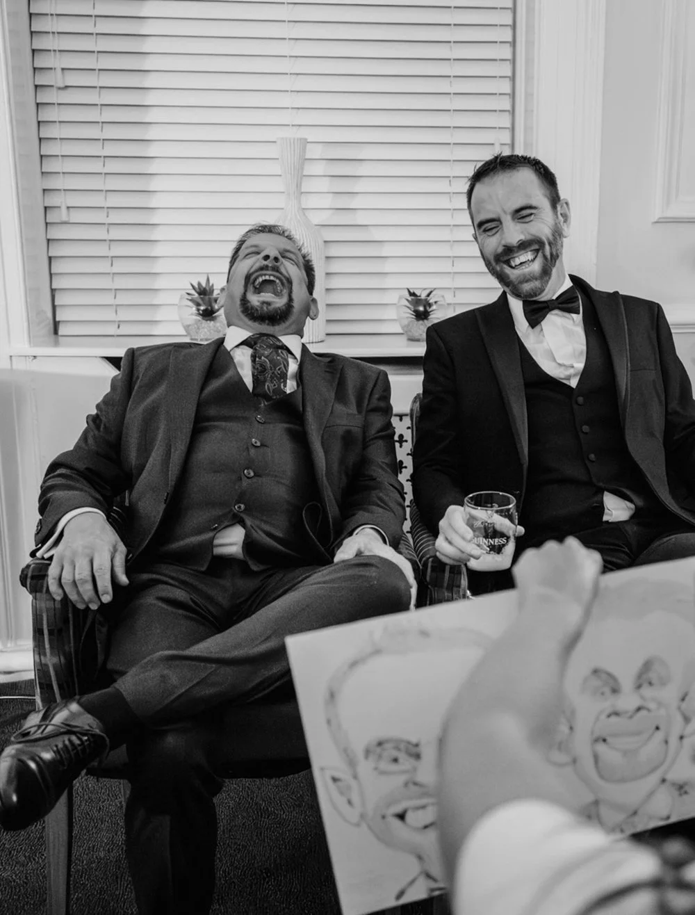 Two men in formal suits sitting and laughing, one holding a glass of Guinness beer, while someone draws caricatures of them in the foreground.