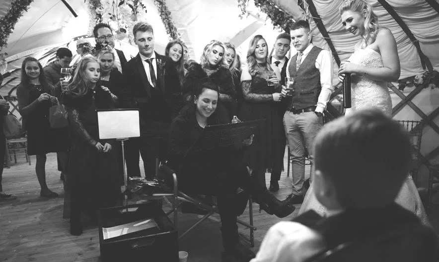 A group of people dressed in formal attire gathered around a woman drawing caricatures during a wedding or special event, inside a decorated tent.