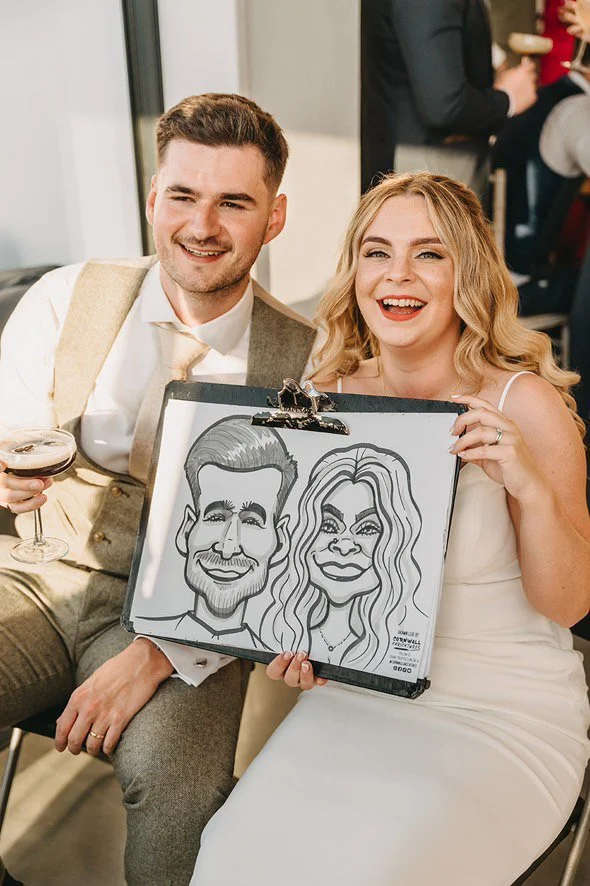 Two smiling people, a man and a woman, holding a caricature drawing of themselves. The woman is in a white dress, and the man is in a light-colored suit. The woman has blonde hair, and the man has short brown hair. The woman is holding the caricature