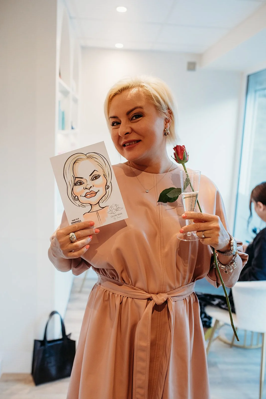 A woman with blonde hair wearing a peach-colored dress holds a caricature portrait, a single red rose in a glass of water, and a glass of champagne, smiling and winking at the camera inside a bright room.