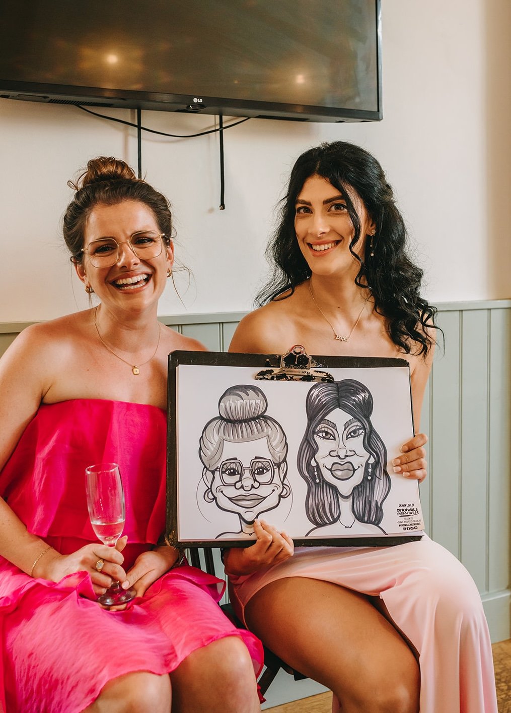 Two women smiling, one in a pink strapless dress holding a glass, the other holding a caricature drawing of both women. The women are seated indoors with a television mounted on the wall above them.