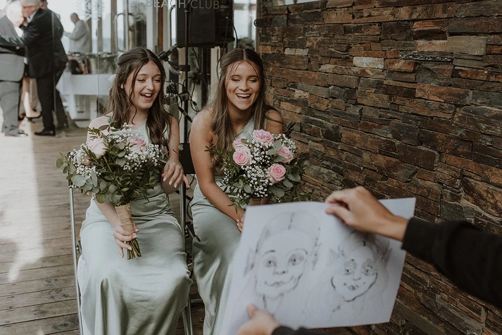 Two women in light-colored dresses sitting on chairs, smiling and holding bouquets of flowers, while an artist sketches their portraits on a canvas.