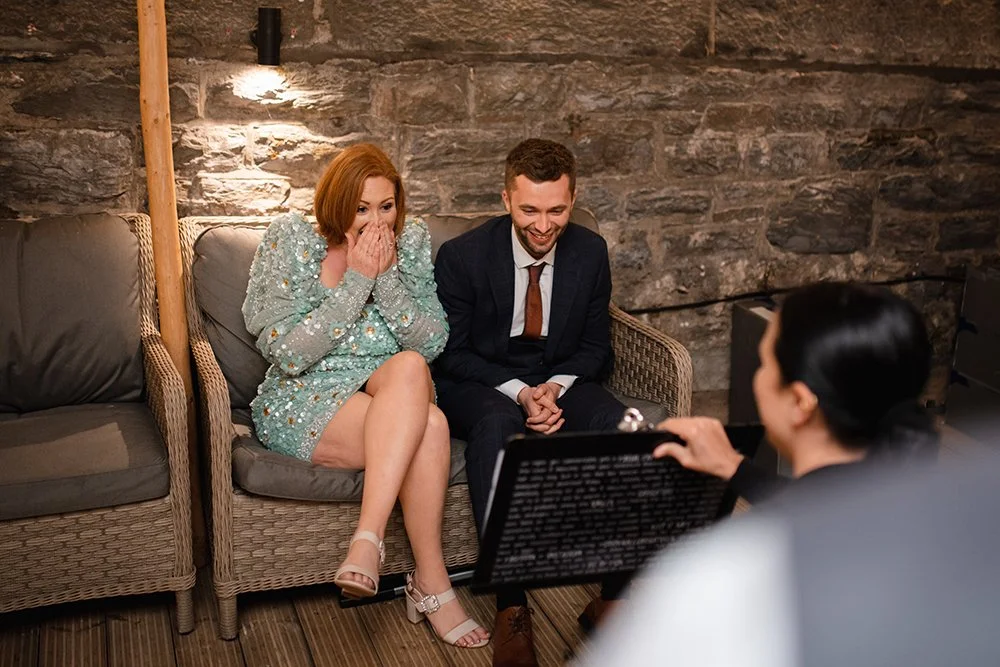 A woman with red hair in a floral dress is surprised and covering her mouth with her hands while sitting on a wicker sofa next to a man in a suit. They are sitting across from a caricature artist in a cozy, rustic room with stone walls.