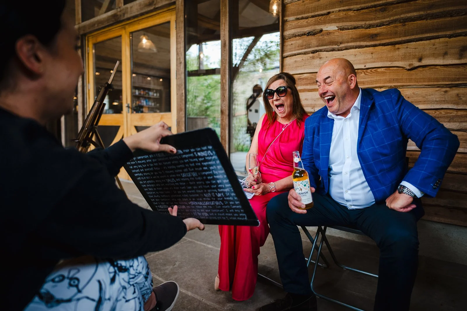 A woman in sunglasses and a pink dress and a man in a blue blazer and white shirt laughing while sitting outdoors, as a live wedding illustrator reveals their live caricature portrait.