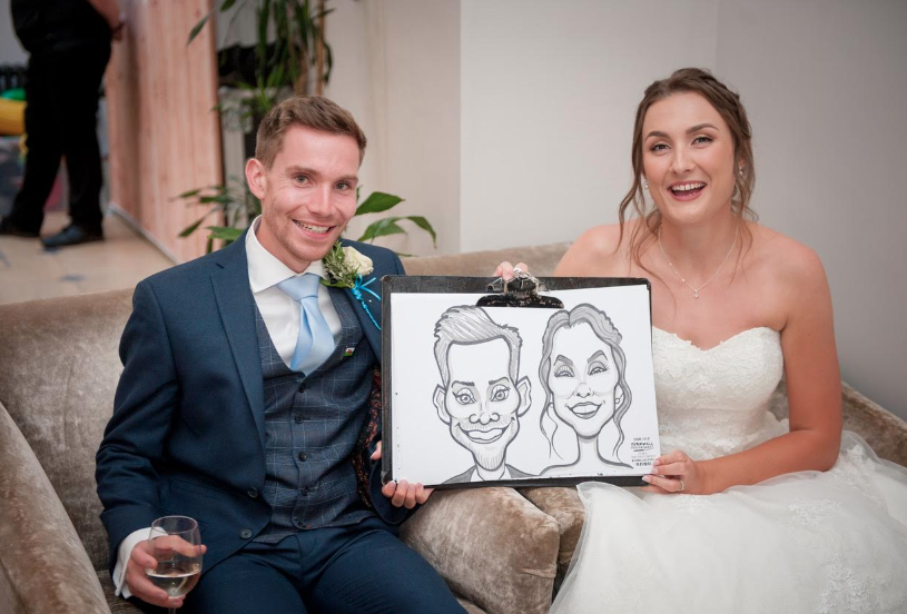 A newlywed couple sitting on a beige sofa, holding a caricature drawing of themselves, smiling at the camera, with a glass of champagne in the groom's hand.
