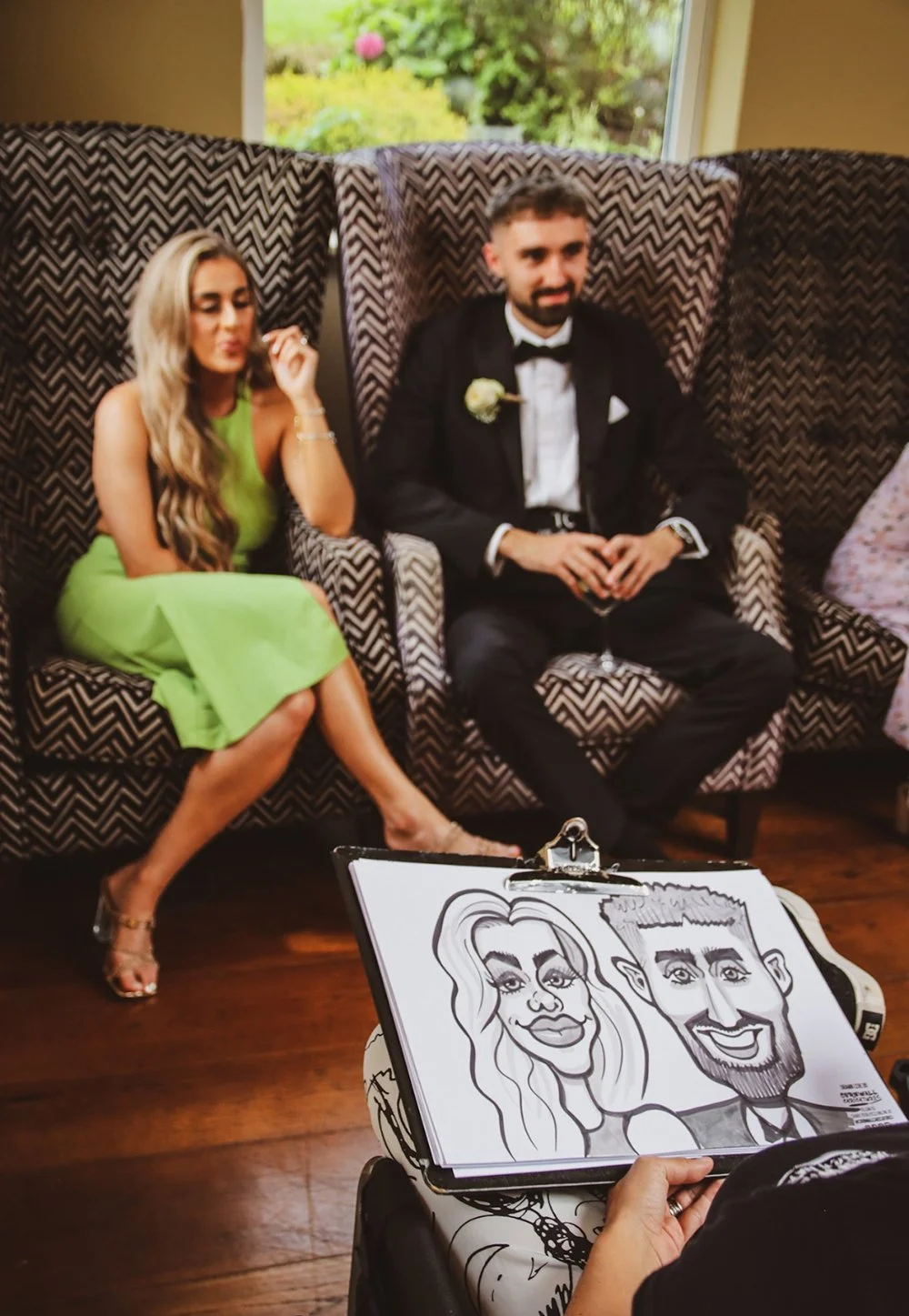 A woman in a bright green dress and a man in a black tuxedo sit on a patterned sofa, getting a caricature portrait drawn of them. The caricature depicts their exaggerated facial features.