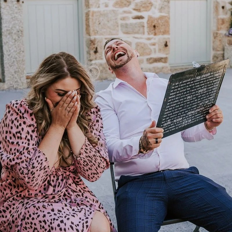 A man and woman sitting outdoors at a wedding reception, laughing and smiling at their caricature illustration; the woman has her hands over her face and the man is holding a clipboard containing his live caricature portrait, looking joyful.