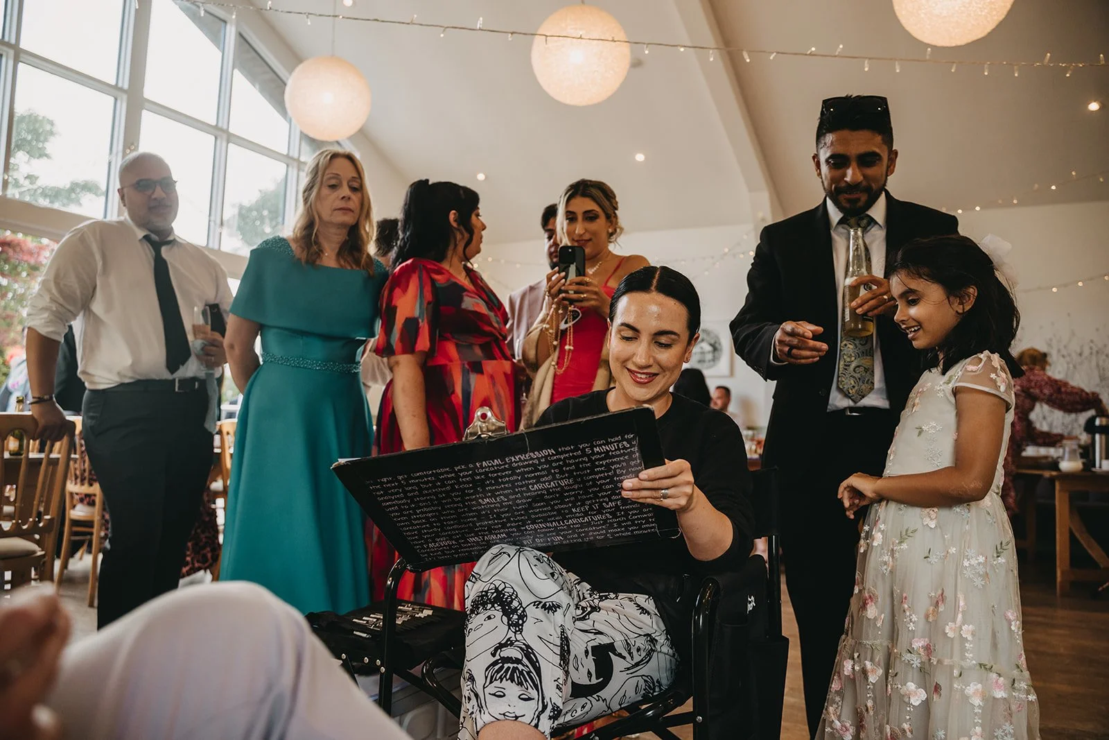 Group of people gathered at a celebration, with a woman in a chair drawing caricatures and others standing around her, smiling and interacting.