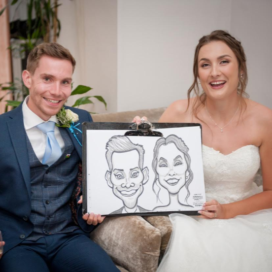 A newlywed couple in wedding attire smiling and sitting on a couch, holding a caricature drawing of themselves.