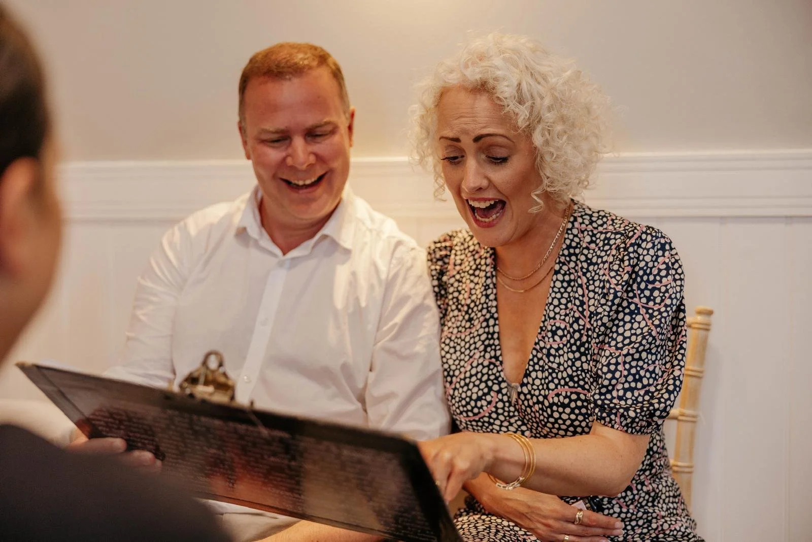 A woman and a middle-aged man are sitting together, smiling and looking at a caricature, enjoying a joyful moment indoors.