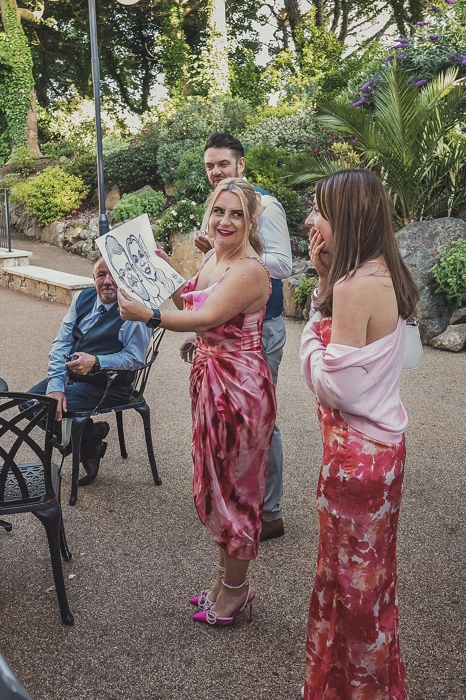 A group of four adults, three women and one man, are standing outdoors in a garden with lush greenery and rocks. The woman in the center is smiling and holding up a caricature drawing of herself, while the other woman is laughing and appears surprise