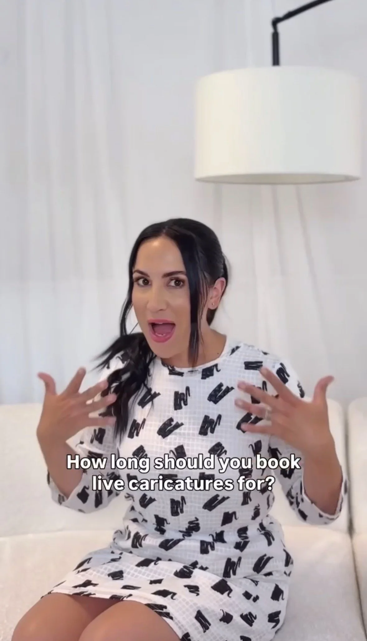 A woman with long dark hair, wearing a black and white patterned dress, is sitting on a white couch in a bright room. She has an expressive facial expression and is gesturing with her hands. There is a white lampshade hanging from a lamp on the wall behind her. Subtitles on the screen read: "How long should you book live caricatures for?"