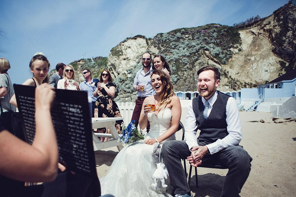 A group of people on a beach during a wedding ceremony or celebration. A bride in a wedding dress and a groom in a suit are seated, laughing and holding drinks, while guests stand behind them, some taking photos.