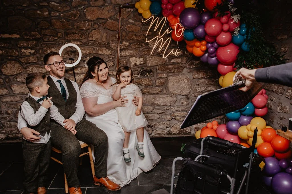A family sitting against a stone wall during a wedding celebration, seeing their caricature revealed, with colourful balloons and 'Mr & Mrs' sign behind them.