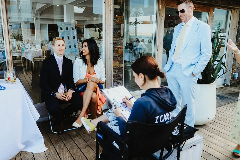 A woman in a patterned dress sits next to a man in a dark suit, both smiling as they talk. A woman with a clipboard and pen in the foreground. A man in a light suit and sunglasses stands near them, smiling. The setting appears to be a patio or outdoo